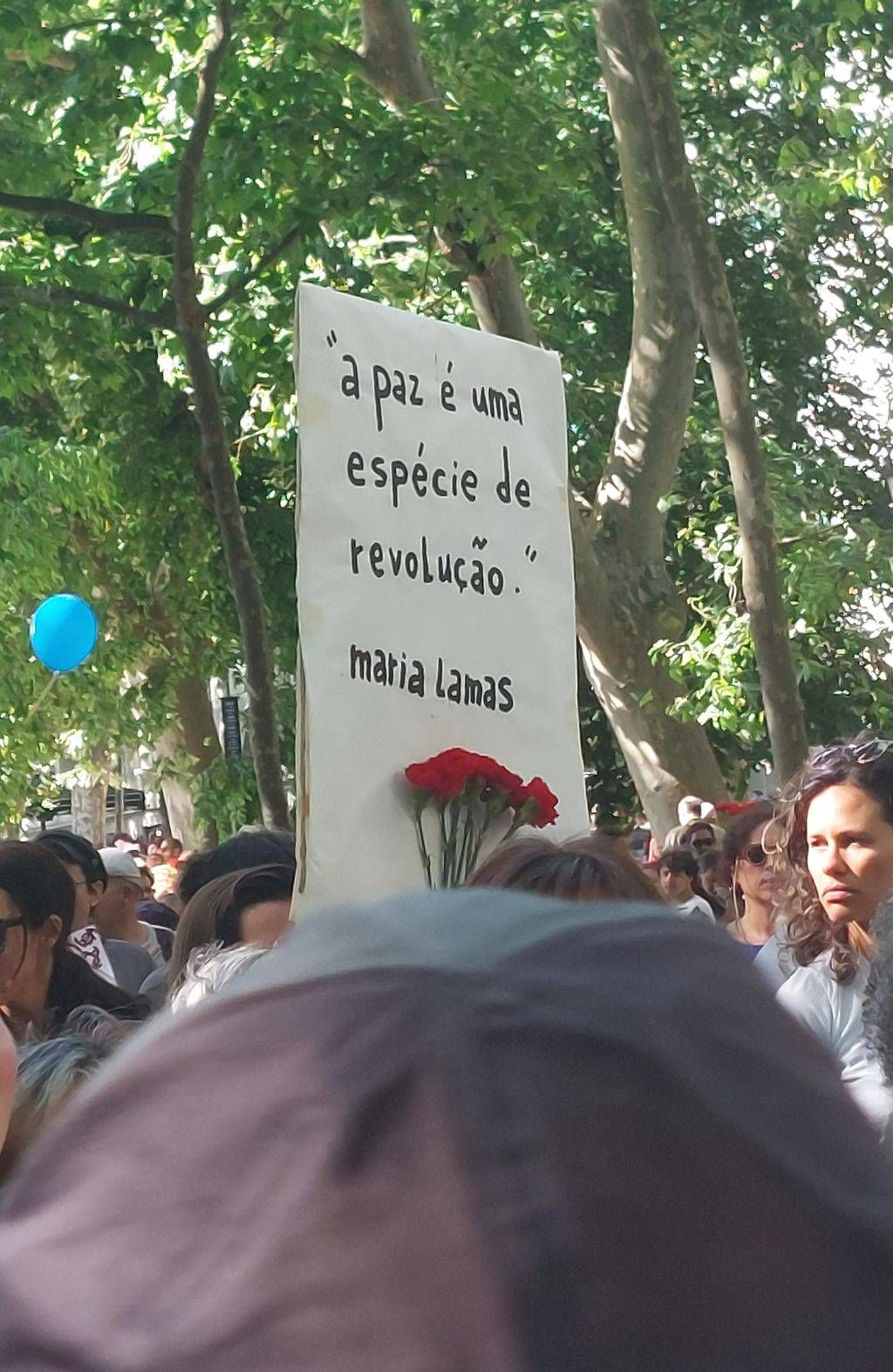 Um cartaz é empunhado no meio da manifestação popular com os dizeres "A paz é uma espécie de revolução" de Maria Lamas. Inclui cravo.