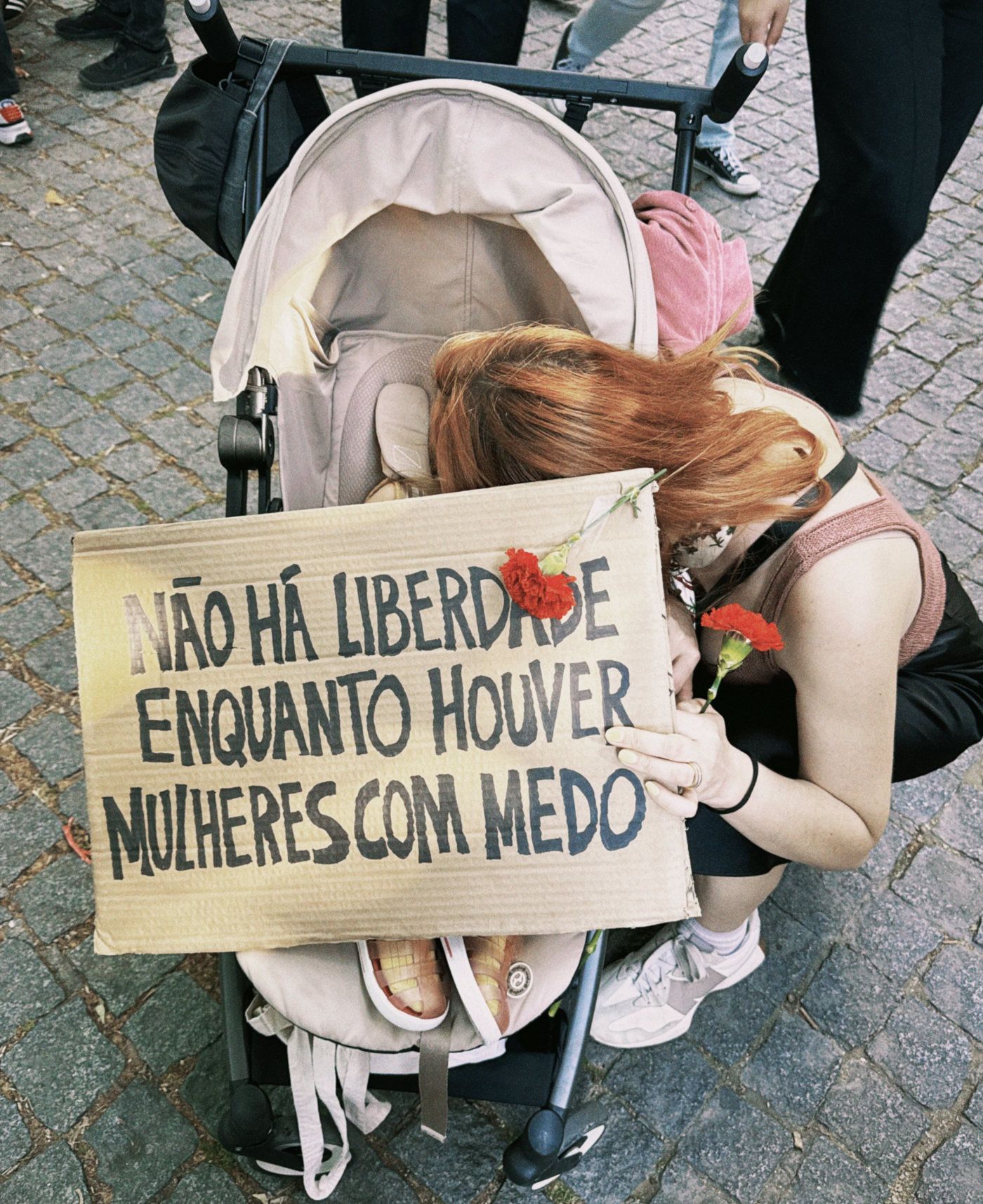 Um mulher na rua aninha-se atrás de um cartaz que diz "Não há Liberdade enquanto houver mulheres com medo", ao lado de um carrinho de bebé.