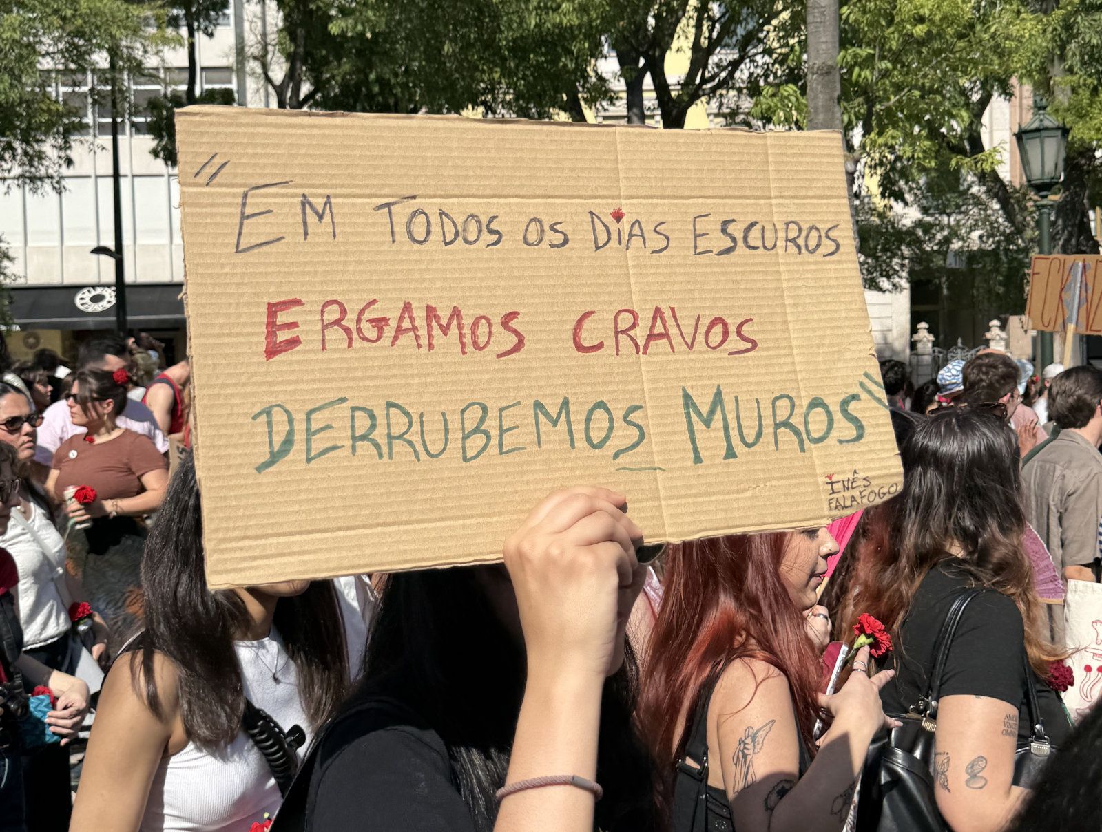 Uma entre outras mãos de jovens raparigas segura um cartaz que diz "Em todos os dias escuros Ergamos cravos, derrubemos muros"