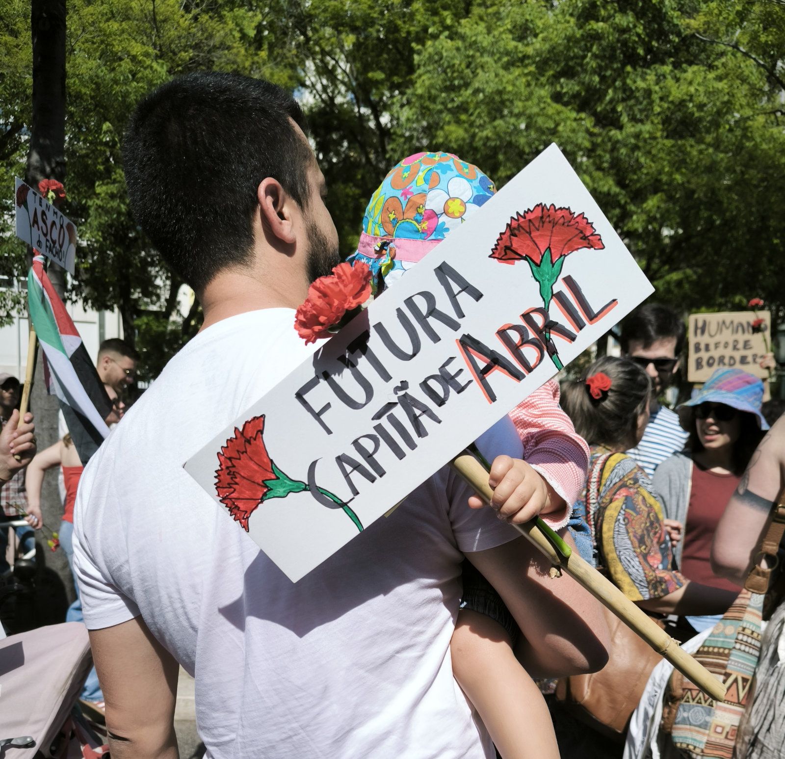Um pai segura um filha ao colo que tem um cartaz com um cravo e as palavras "Futura capitã de Abril"