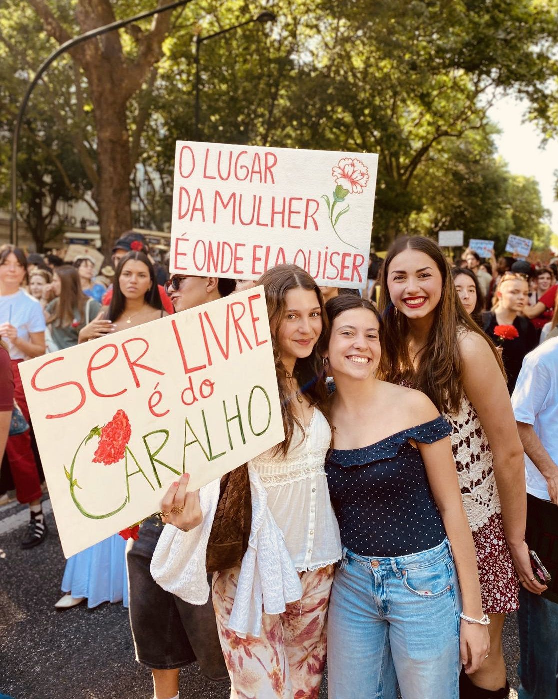 Três mulheres seguram dois cartazes num avenida. Cartazes rezam "O lugar da mulher é onde ela quiser" e "Ser livre é do Caralho"