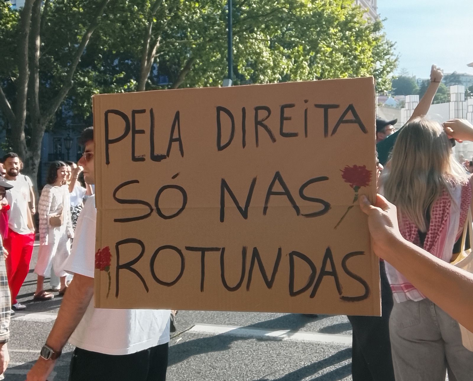 Um mão segura um cartaz no meio da multidão que diz "Pela direita, só nas rotundas"